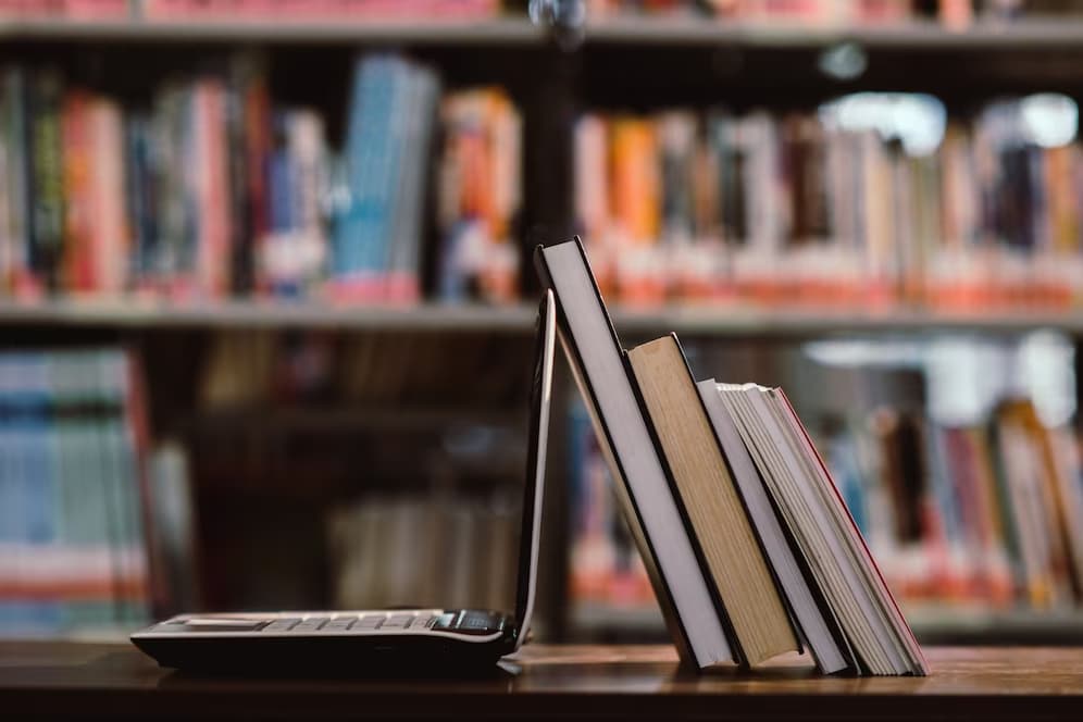 Imagem de um computador com livros em uma biblioteca, para remeter ao edital de conservação do Rio de Janeiro