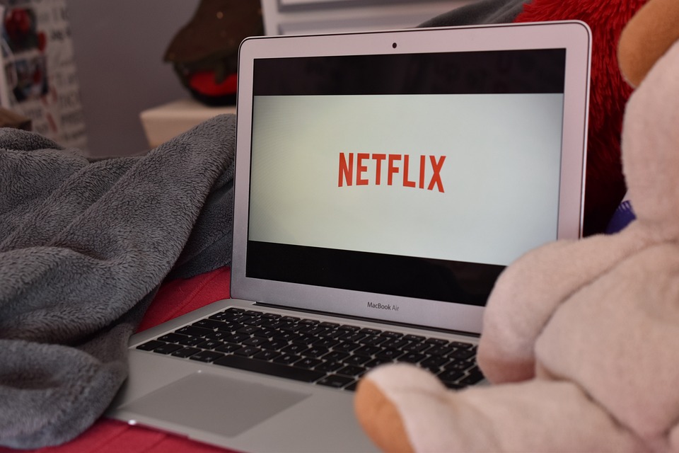 Notebook sobre cama com Netflix na tela, ao lado de lençol e ursinho de pelúcia.